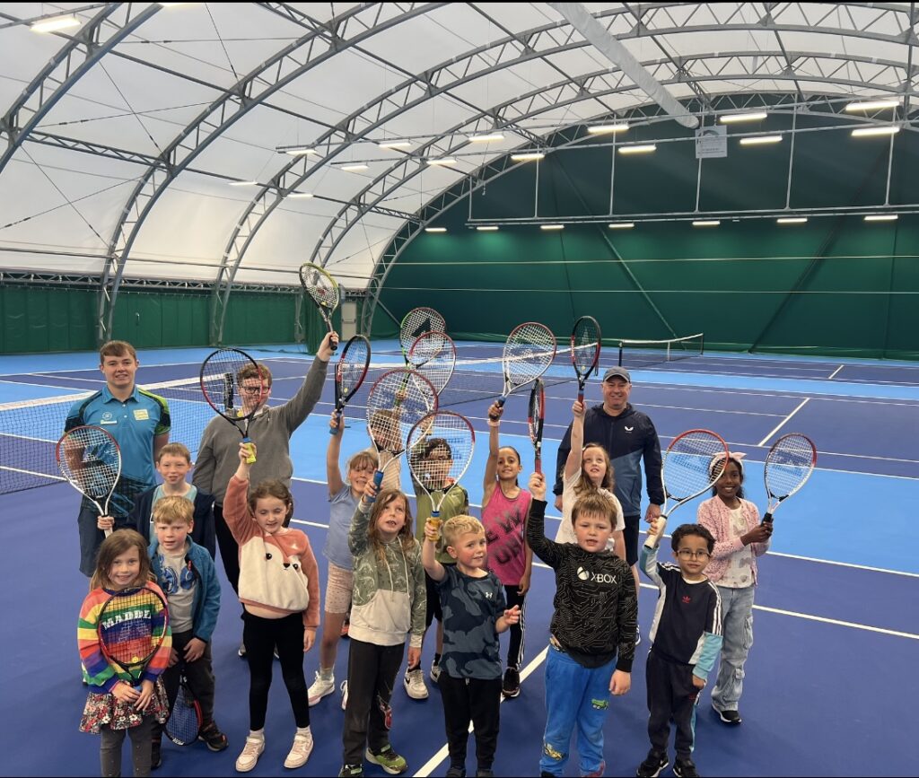 A group of young kids holding up their tennis rackets for a photo - some staff members in the back