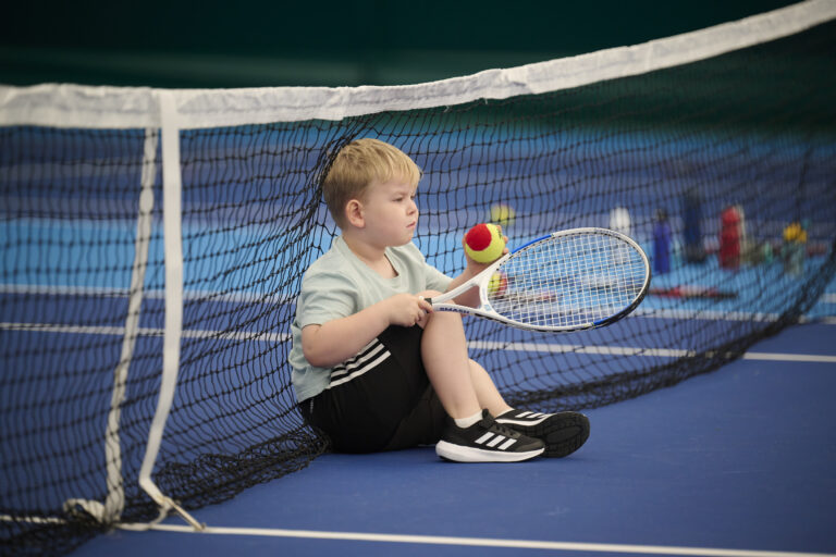 A picture of a young boy sitting against the tennis net holding a tennis racket and ball
