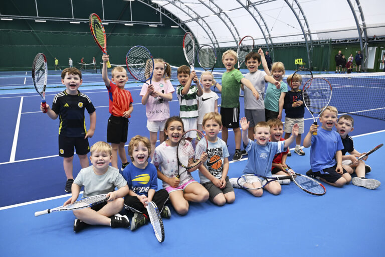 A large group of kids looking really happy and holding their tennis rackets in the air at moray sports centre